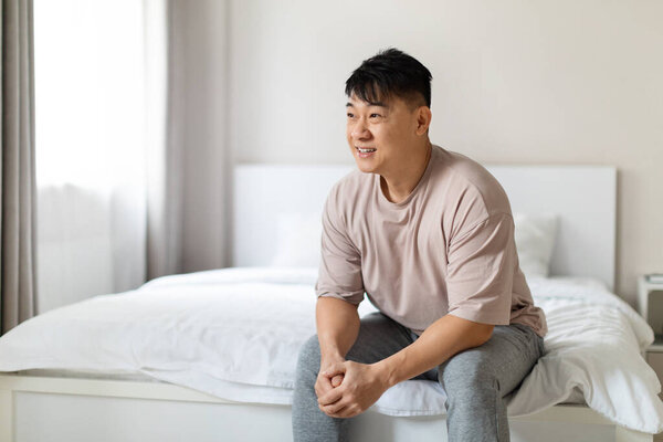 Happy middle aged asian man enjoying sunny morning, sitting on comfy bed in white cozy bedroom after waking up, feeling excited and well-rested, looking at copy space and smiling