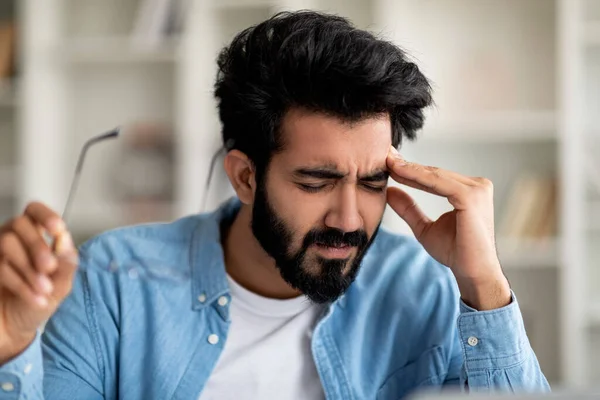 疲倦的印度年轻人在家里患有急性头痛 有病的东方男性自由职业者摘下眼镜擦拭圣殿 千禧年的男人下班后感到精疲力竭 — 图库照片
