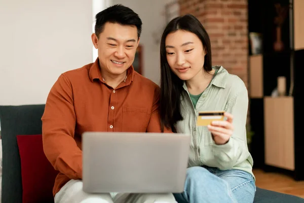 微笑的韩国中年男子和年轻女子使用信用卡和笔记本电脑 坐在客厅的沙发上 检查财务账户 应用银行业务 购物狂享受销售 — 图库照片
