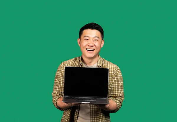 Happy asian middle aged man showing laptop computer blank screen, recommending great website, standing on green studio background, mockup. Online service advertisement