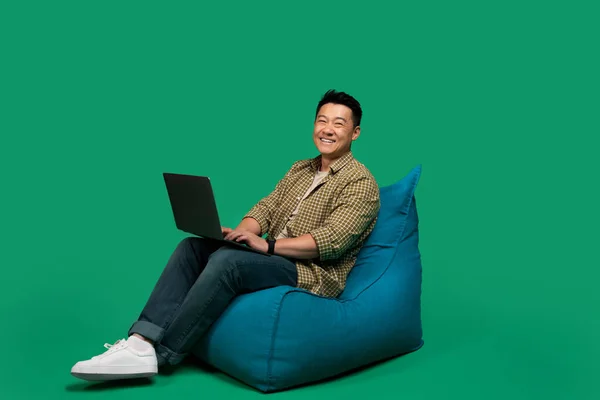 Happy asian middle aged man using laptop, working online while sitting in beanbag chair over green studio background, free space. Male freelancer browsing internet and smiling