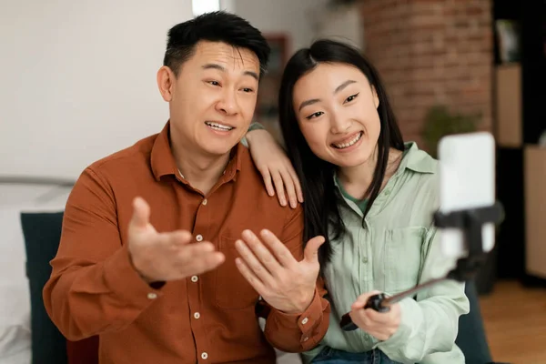 幸せなアジアの男性と女性は自宅でソファに座って 携帯電話を使用して 家族とビデオ通話をしたり 三脚にスマートフォンのウェブカメラで話したりジェスチャーしたりします — ストック写真