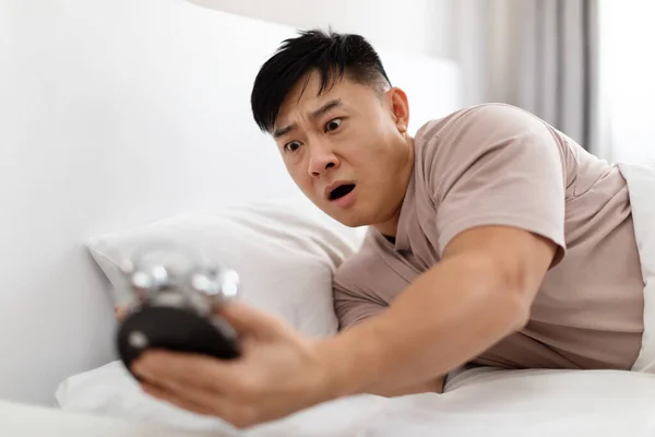 Shocked upset overslept handsome middle aged asian man wearing pajamas lying in bed at home, looking at alarm clock, late for flight or job, need to hurry up, copy space