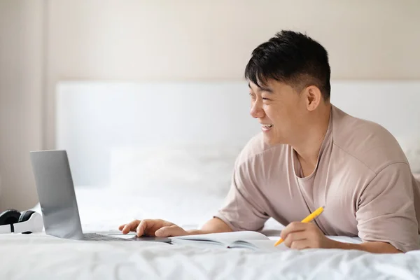快乐的中年亚裔男子穿着睡衣在家里学习 参加网络研讨会或网上培训 在床上使用笔记本电脑 记笔记 复制空间 网上教育 — 图库照片