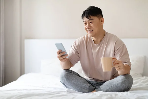 快乐而又正派的中年男子穿着睡衣坐在床上 使用智能手机和咖啡 阅读新闻 醒来后查看社交媒体 复制空间 — 图库照片