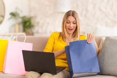Bin yıllık neşeli, beyaz, sarışın bayan alışveriş meraklısı. Kanepede dizüstü bilgisayar kullanarak alışveriş yapıyor. Oturma odasında online alışverişin keyfini çıkarıyor. Paketleri açmak, büyük indirim, nakit para, evde sipariş vermek.