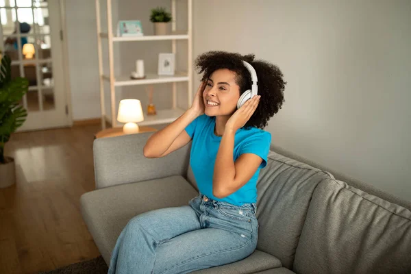 ワイヤレスヘッドフォンで幸せな千年のアフリカ系アメリカ人の巻き女性は リビングルームのインテリアでソファの上で無料の時間をお楽しみください 休息とリラックス 学習とレッスンのオーディオアプリ — ストック写真