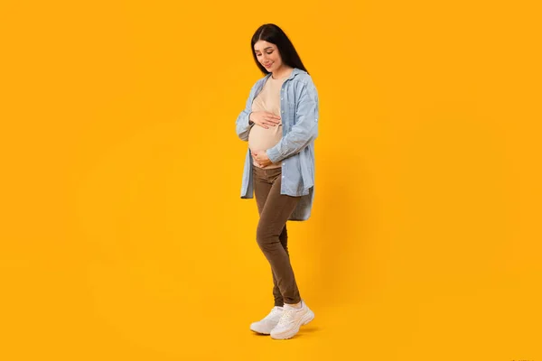 健康怀孕概念 年轻的孕妇摸着她的腹部 看着它 孕妇站在黄色的背景上 全身而退 复制空间 — 图库照片