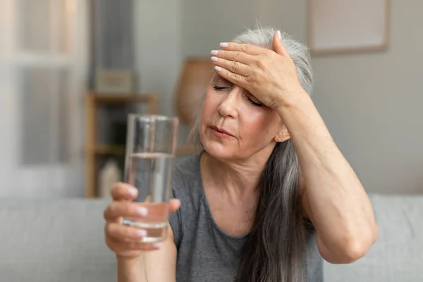 Unhappy sad caucasian old lady hold glass of water and touching forehead, suffering from fever, menopause in living room interior. Health problems, treatment of disease, high pressure and migraine