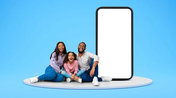 Laughing excited millennial black man, woman and little girl sit on floor with huge phone with empty screen on blue studio background. Technology, app and website for family