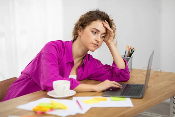 退屈な仕事だ 若いフリーランスの女性が自宅のノートパソコンで働いていて 机の上に座っている女性をパソコンで動揺させ 退屈そうな顔の表情で画面を見て 単調な仕事に疲れている — ストック写真