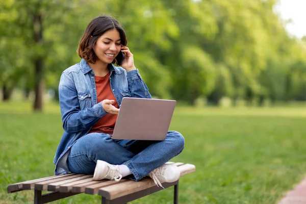 若いアラブ人女性が携帯電話で話しているとノートパソコンの屋外を使用して笑顔の肖像 キャンパスでベンチに座っている間 幸せな中東の女性は 電話の会話を楽しみ ノートパソコンで作業 — ストック写真