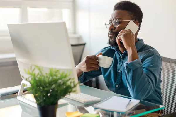 Remote business. African american male ceo talking on cellphone and using computer, male having corporate phone conversation call and drinking coffee
