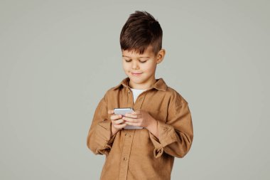 Gri arka planda izole edilmiş telefon oyununda oynayan 6 yaşındaki gülümseyen beyaz çocuk. İnceleme uygulaması, cihazla boş zaman eğlencesi, çocukluk ve çevrimiçi eğitim, alet bağımlılığı