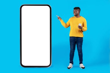 Mobil Teklif. Gülümseyen genç siyah adam, Beyaz Ekranlı Büyük Boş Akıllı Telefonu işaret ediyor, Mutlu Afro-Amerikan Adam Başvuru veya Web Sitesi, Mavi Arkaplan 'ın üzerinde duruyor,