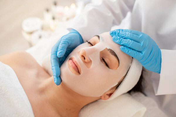 Modern Cosmetology Concept. Beautician Doctor Applying Sheet Mask On Face Of Young Female, Attractive Female Getting Moisturising Skincare Treatment In Luxury Spa Salon, Closeup Shot