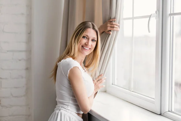 Happy Millennial Caucasian Blonde Woman Domestic Clothes Pulls Back Curtain — Stock Photo, Image