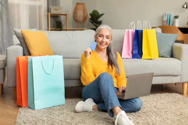 Smiling caucasian old lady with a lot of bags from store sits on floor enjoys online shopping, recommends credit card, use laptop in living room interior. Huge sale, banking and finance for order