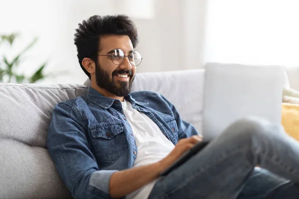 Smiling Handsome Indian Man Using Laptop While Sitting On Couch In Living Room At Home, Young Eastern Male Freelancer Working On Computer Or Communicating Online, Closeup Shot With Free Space