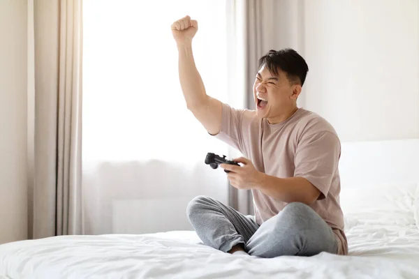 兴奋而快乐的中年男子 穿着睡衣或家居服 坐在床上 拿着控制杆 举手表决 玩电子游戏 复制空间 — 图库照片