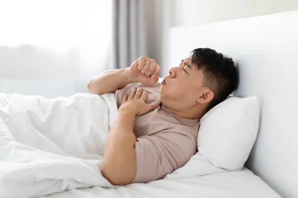 ベッドに横たわって咳をし 彼の胸 自宅のインテリア サイドビュー コピースペースに触れるパジャマを着て不健康な中年のアジア人男性 肺癌肺炎喫煙者咳 — ストック写真