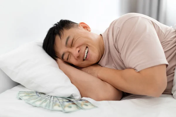 Smiling chinese guy enjoying his sleep while keeping money dollar cash under pillow, middle aged asian man saving money at home, closeup. Financial literacy, savings concept