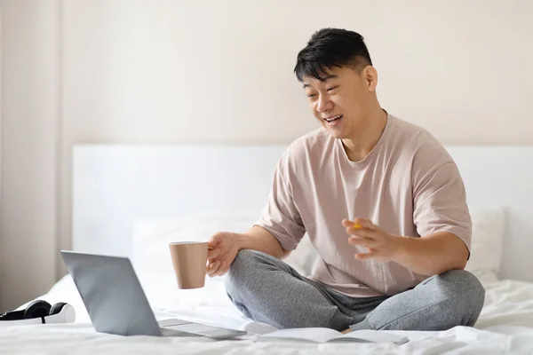 快乐而英俊的中年男人 穿着睡衣坐在床上或宾馆里 用笔记本电脑 拿着咖啡杯 看着电脑屏幕 微笑着 摆出姿势 进行视频聊天 复制空间 — 图库照片