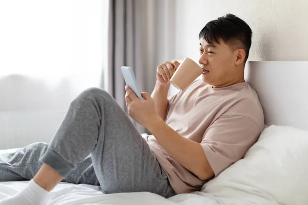 在家里躺在床上 用手机 喝咖啡 穿睡衣 在网上看新闻 查看邮件 看边景 复制空间的中国中年男子 — 图库照片