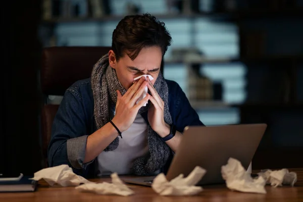 Unhealthy sick young man with scarf around his neck employee working late at office, guy sitting at workdesk, looking at laptop screen and sneezing, using napkin, got flu, coronavirus, copy space