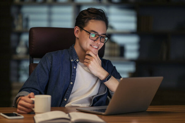 Happy attractive young businessman working overtime alone at his desk in office late at night, wearing eyeglasses, looking at computer screen and smiling, have good news, drinking coffee, copy space