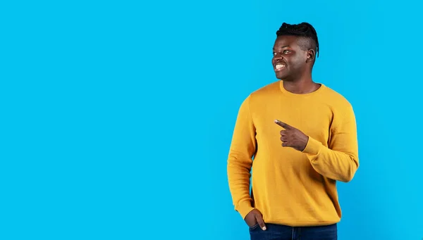 Handsome Black Man Pointing Aside At Copy Space With Hand, Smiling Young African American Guy Demonstrating Free Place For Advertisement While Posing Over Blue Studio Background, Panorama