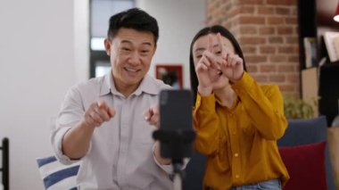 Online içerik. Pozitif Asyalı erkek ve kadın akıllı telefondan komik moda videolar kaydediyor. Cihaza kalp jesti ve kahkaha, telefon tripoda, seçici odaklanma, yavaş çekim.