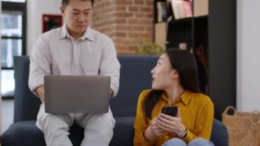 Çevrimiçi kazandı. Genç Asyalı kadın akıllı telefondan internette sörf yapıyor ve kazanan mesajı alıyor, dizüstü bilgisayarda çalışan kocaya telefonu gösteriyor, birlikte başarının tadını çıkarıyor, çekimin izini sürüyor, yavaş çekim, serbest alan.