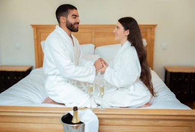 Romantik Teklif. Mutlu çift yatakta el ele tutuşuyor, otel odasında şampanyayla buluşuyor, bornoz giyiyorlar. Aşk İlişkileri Konsepti. Yan Görünüm