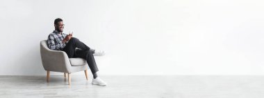 Neşeli başarılı Afro-Amerikan milenyum işadamı sandalyede oturuyor ve ofisteki boş alanda beyaz duvarda akıllı telefonuyla yazı yazıyor. İş için aygıtla iş, yeni normal