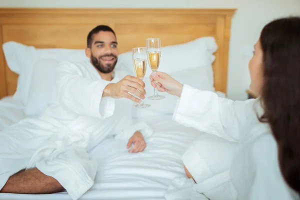 ロマンチックな休暇 幸せなカップルは 豪華なホテルスイートの屋内でベッドに寝そべっている日付を持ち 白いバスローブを着たスパークリングワインを飲むグラスをクリックしてください 選択的フォーカス — ストック写真