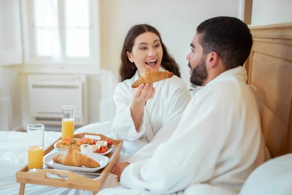 ホテルロマンス 幸せな妻の餌夫はベッドで朝食を食べながらクロワッサンを与えます 寝室の屋内で食品とテーブルトレイに座ります 2人で休暇を楽しむ 選択的フォーカス — ストック写真