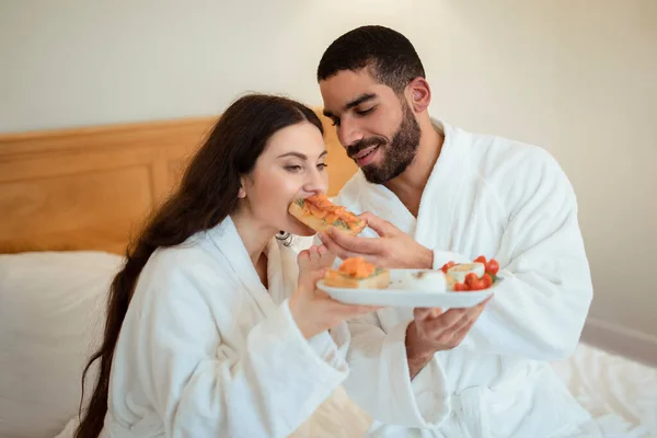 幸せな夫は 家庭で近代的なベッドルームに座ってベッドでロマンチックな朝食を持っている彼女にサンドイッチを与える妻を給餌 朝は白いバスローブを着たカップル — ストック写真