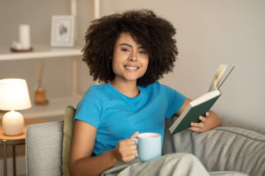 Mutlu binlerce yıllık Afro-Amerikan kıvırcık kadın kitap okuyor, rahatlıyor ve oturma odasındaki kanepede çay içiyor. Dinlen, akşamları evde tek başına ders çalış.