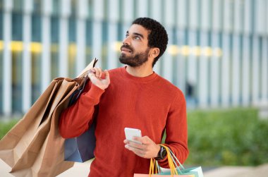 Mobil Alışveriş. Orta Doğulu Alıcı, elinde Akıllı Telefon ve Kağıt Alışveriş Torbası tutuyordu. Dışarıdaki modern alışveriş merkezinin yanında poz veriyordu. Erkek Alışveriş Komitesi Reklamcılığı Büyük Satış Teklifi