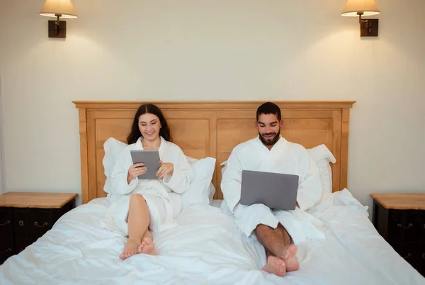 Happy Spouses Browsing Internet On Computers Using Laptop And Digital Tablet Sitting In Bed In Hotel Room Indoors, Wearing Bathrobes. Freelance Career And Internet Technology Concept