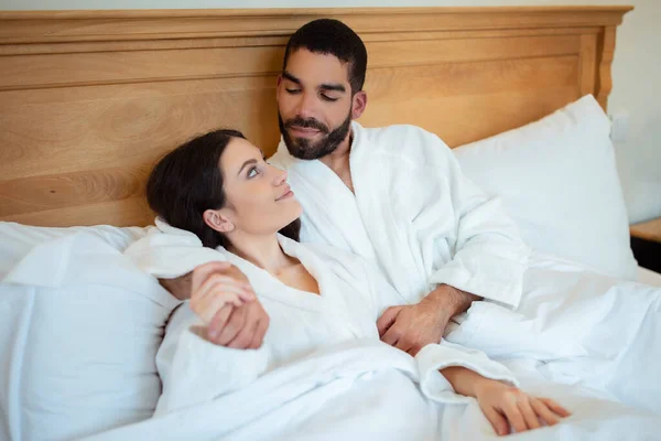 寝室の屋内にベッドで寝そべっていることを受け入れる愛の多様なカップル 白いバスローブを着て 休暇中にホテルスイートでお互いをリラックスして見て配偶者 ロマンチックな新婚旅行 — ストック写真
