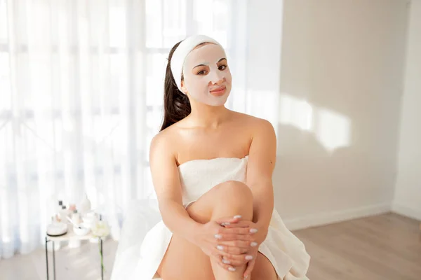 Self-Care Concept. Portrait Of Beautiful Woman With Sheet Mask On Face Sitting Wrapped In Towel After Bath Inddors, Attractive Female Enjoying Home Beauty Treatments And Skincare, Copy Space