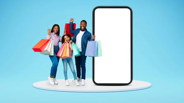 Glad millennial african american mom, dad and small girl hold shopping bags, family rejoice purchase, near huge smartphone with empty screen on stage on blue studio background. Sale and discounts