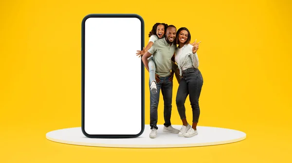 小さな娘を持つ幸せなアフリカ系アメリカ人の家族は 黄色の背景 スタジオでステージ上に空の画面を持つ大きな携帯電話と一緒に楽しみを持っています 親と子 アプリ — ストック写真