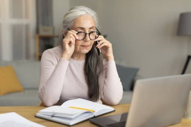 Oturma odasındaki bilgisayarlı telefondan arayan, gözlüklü, oldukça meşgul, beyaz, yaşlı bir kadın. Teknolojiyle iletişim, iş ve iş hayatı, Covid-19 'a olan sosyal uzaklık