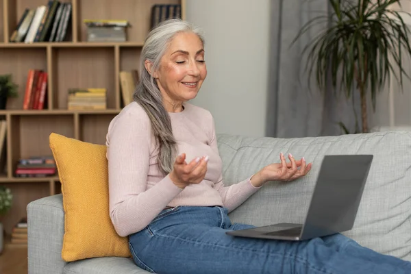 快乐的高加索老年女性坐在沙发上 有视频通话 看电脑 在客厅里摆姿势 在家里举行远程会议 技术交流 聊天和社交距离 — 图库照片