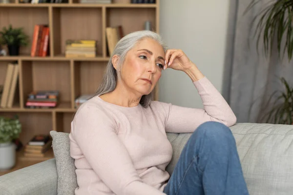 Sad tired depressed caucasian senior woman sitting on sofa, suffering from menopause, migraine, pressure and stress in living room interior. Reaction to bad news, health problems and emotions at home