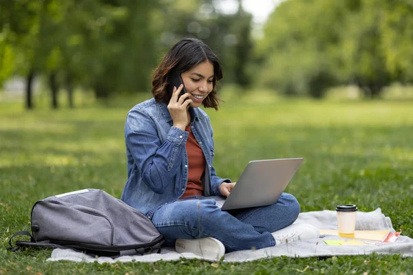 Smiling Young Arab Woman Using Laptop And Cellphone While Relaxing Outdoors, Happy Middle Eastern Female Student Using Computer And Talking On Mobile Phone While Sitting In City Park, Copy Space