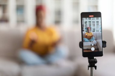 Cep telefonunun seçici odak noktası kırmızı türban vlogger 'lı sevimli Afrika kökenli Amerikalı bir kadının sosyal medyada takipçileri için yayınlanması, konuşması, jestleri, bulanık geçmişi.